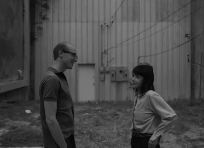 A bespectacled man in a polo and a dark-haired woman in a white blouse stare at each other in front of an industrial-style building.