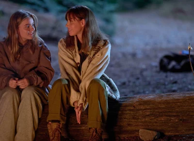 Two women sit on a log, looking at each other.