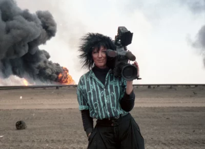 Margaret Moth stands in front of a distant explosion with a camera on her shoulder.