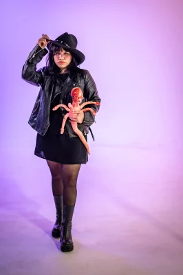 Aura Martinez Sandoval holds a gore-y prop, tipping their cowboy hat and wearing a black leather jacket against a purple background. 