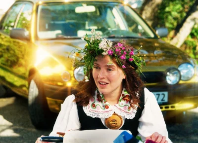 A young woman in a flower crown stands in disbelief in front of a shining gold-plated car.