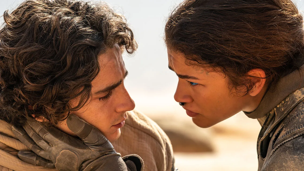 A young woman caresses a young mans face while staring into his eyes, they're surrounded by desert.