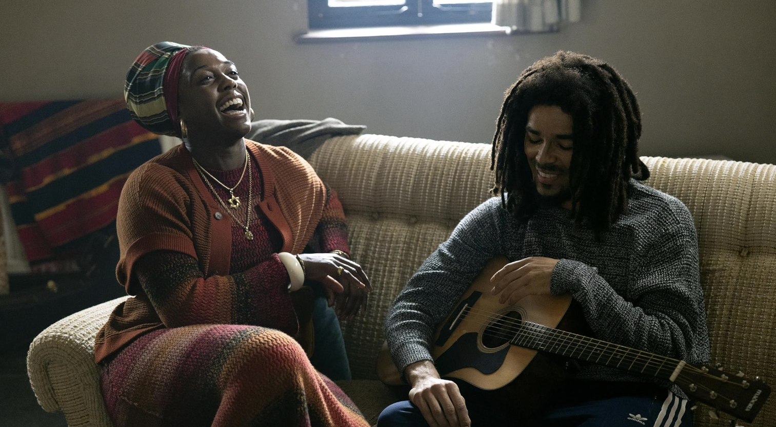 Lashana Lynch as Rita Marley and Kingsley Ben-Adir as Bob Marley laugh together on a couch.