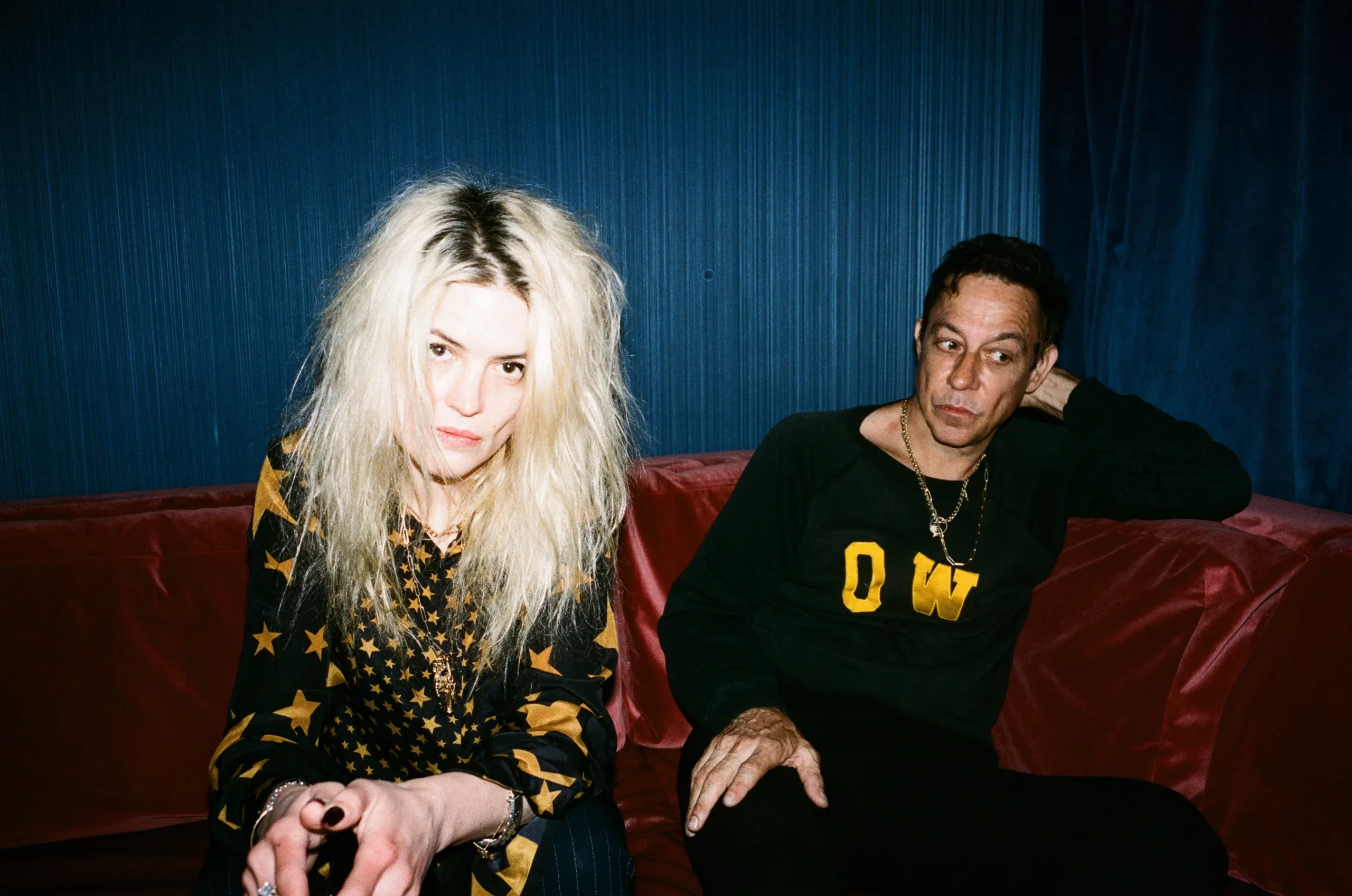 Alison Mosshart and Jamie Hince sit on a red velvet couch with a blue curtain behind them.