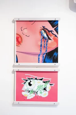 Two pink paintings depicting anime looking eyes hang on a white wall.