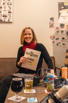 Angie Keeton smiles holding a hymnal with beer in front of her.