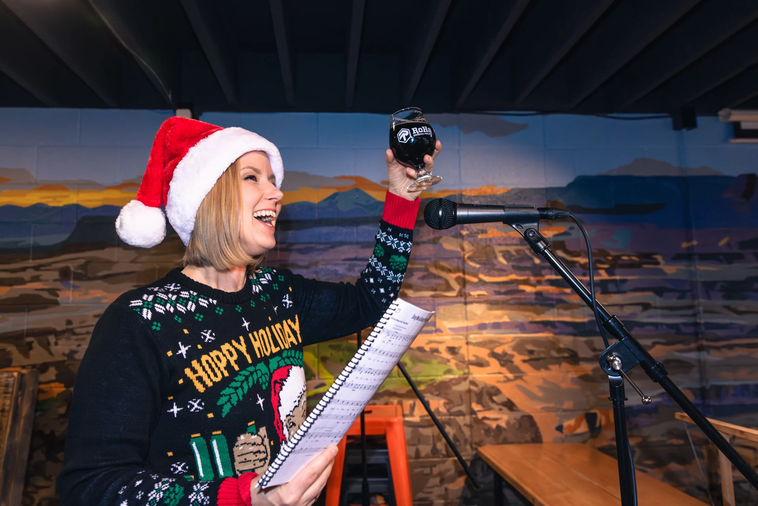 A woman in a Santa hat and an ugly Christmas sweater stands in front of a mic holding sheet music in one hand, a beer raised in the other.