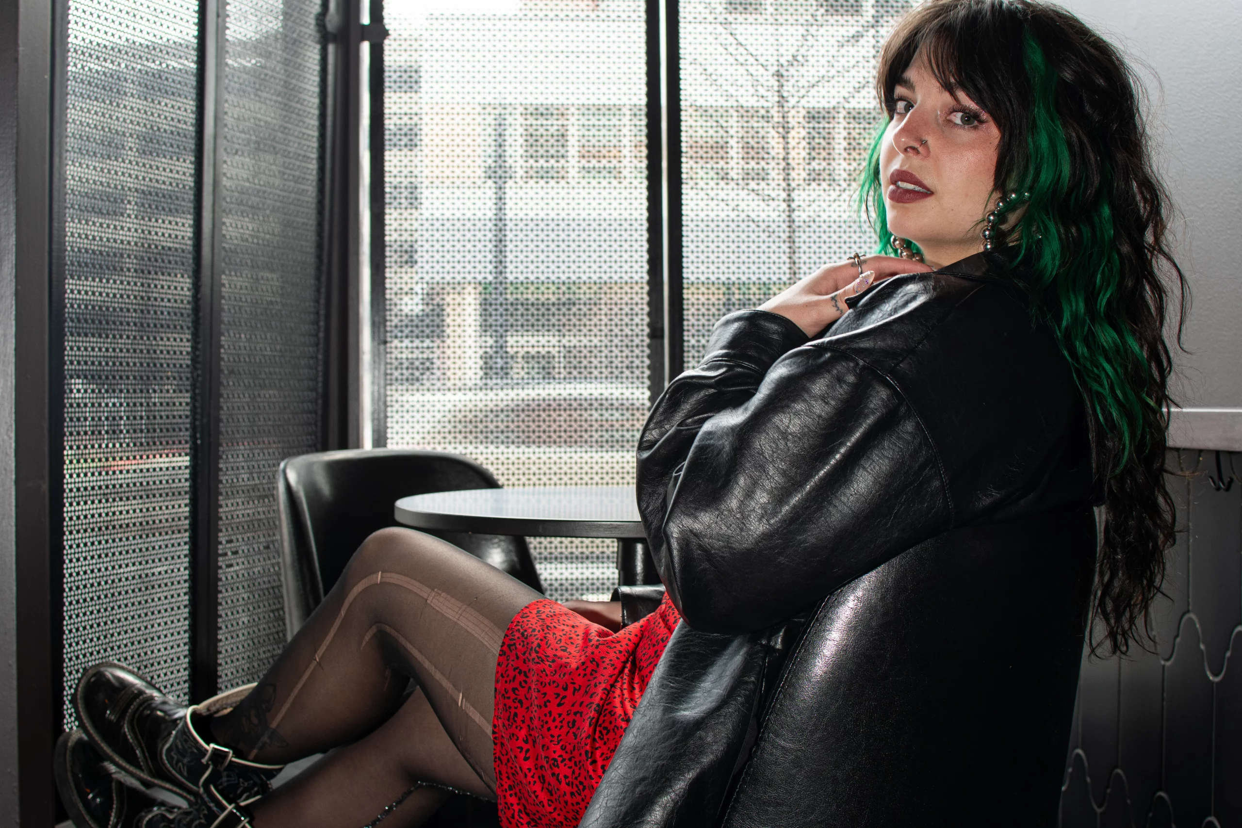 Girl with black hair and cool outfit sits in a chair posing at the camera.