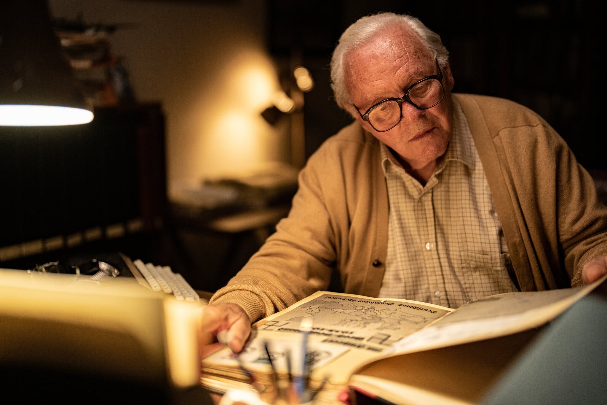 An older bespectacled gentleman surrounded in warm light looks over a large book of text.