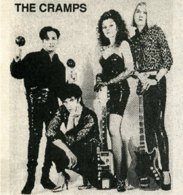A group of four individuals stand posing with various instruments while their band name, The Cramps, appears in black text in the top left corner of their white backdrop. 