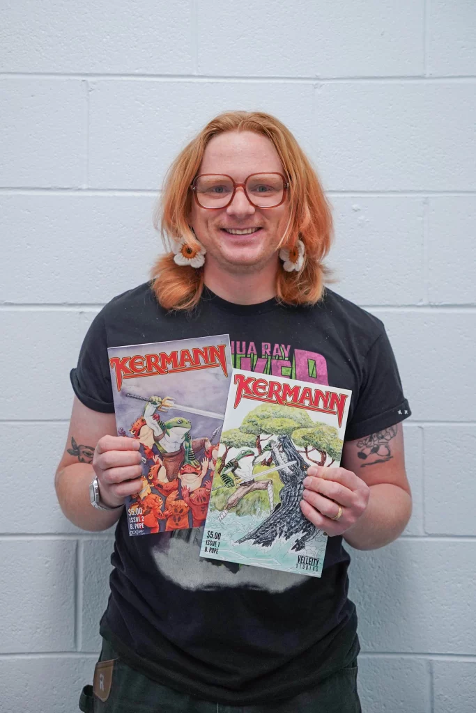A person with shoulder length ginger hair holding comics stands in front of a white wall. They smile into the camera.