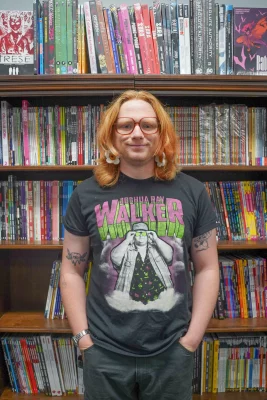A person with shoulder-length ginger hair and big red glasses stands in front of a bookcase full of comic books. 