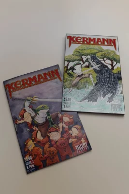 Two comic books featuring frogs holding swords sit on a white backdrop. 