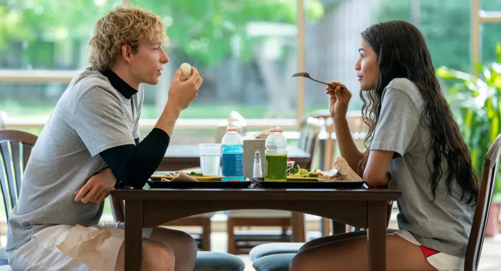 A young blonde man sits across a table from a young girl with long dark hair. Over the table of hard boiled eggs and Gatorade, she points a fork while in conversation with the young man.
