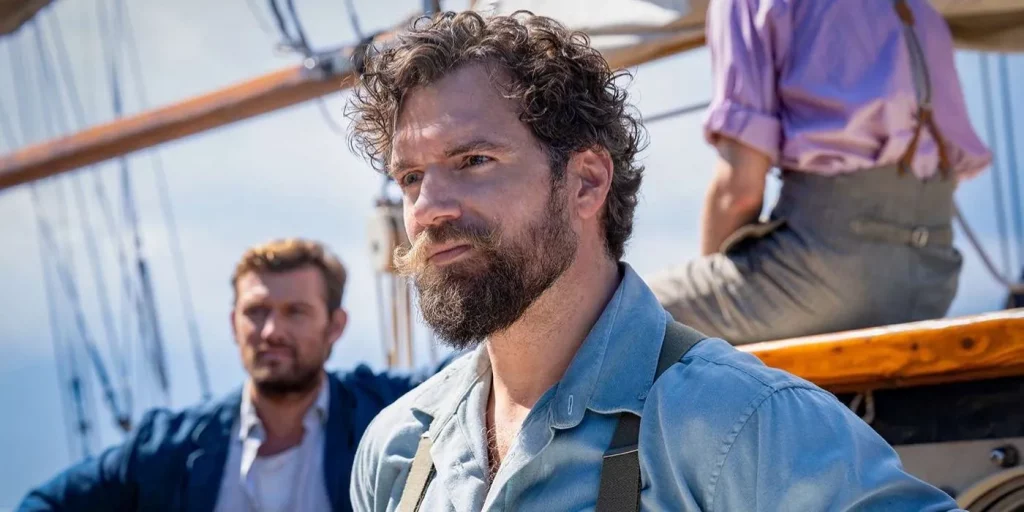 A bearded man with curly hair looks to left of the screen. He appears to be on a ship surrounded by crew.