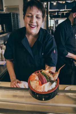A woman in a black chef's uniform smiles as she holds a sushi bowl towards the camera. 