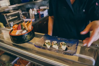 A sushi bowl and a plate of three individual pieces of fish drizzled in sauce sit atop the sushi bar counter.