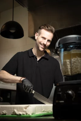 A man in a black collared shirt stands in a kitchen cutting a brick of white vegan cheese. He smiles into a camera, a blender full of ingredients in the foreground.