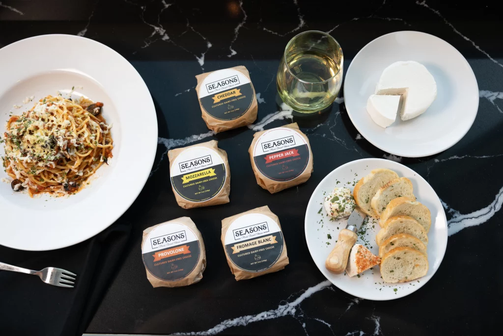 Seasons Cheese five cheese flavors sit atop a black table. They're surrounded by a plate of spaghetti, a plate of sliced bread, and a plate of a wheel of cheese.