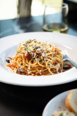 An image of plated spaghetti topped with Seasons vegan cheese atop a black table. A wineglass out of focus in the background.