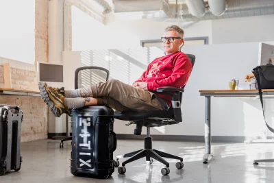 Shon Taylor sitting on a rolling office chair resting his legs on a 24tix suitcase.