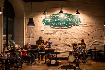 A band of musicians play at Two-Bit Bistro in Ogden, Utah, in front of a wall displaying the restaurant's green logo and sign.