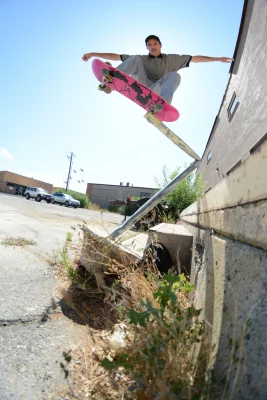An action shot of Landen Fergus skating off the edge of a building on a pink skateboard, the sun glowing behind him.