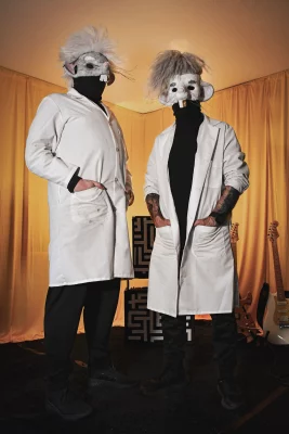 Devin (left) and Josh (right) stand together in labcoats and rat masks with their hands in their pockets.