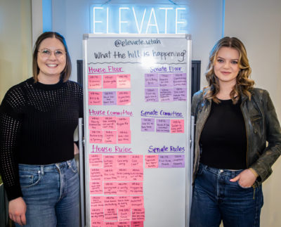 Finlayson and Morgan from Elevate stand in front of a whiteboard covered in informative pink and purple sticky notes. A neon blue "ELEVATE" sign glows overhead.