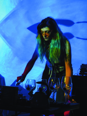 Hal Jean stands in blue-green light in front of a row of water-filled wine glasses, playing music on them.