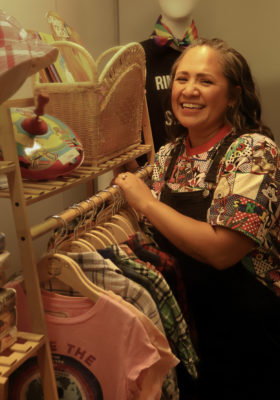 The owner of Bright & Stormy Kids is standing near a rack of clothing in her store.