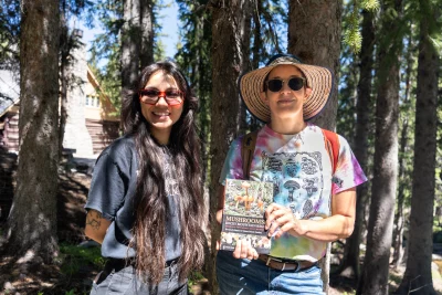 Two women standing in the woods, one is holding a field guide to mushrooms.
