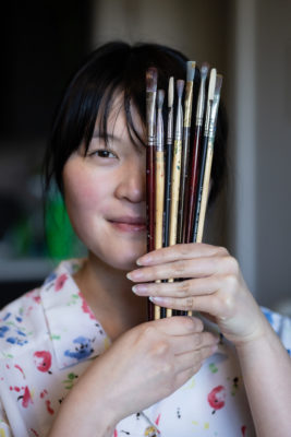 Mercedes Nokyi Ng holding a clutch of paintbrushes over one eye.