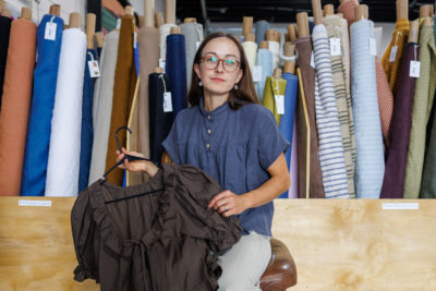 Delaney Dangerfield of Salt Lake Sewciety in front of bolts of fabric, holding a hand-made shirt on a hanger.