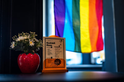 A tabletop cocktail menu at Spritz! with a pride flag hanging in the background.
