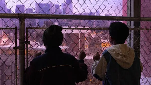 Two young boys look out at a lilac sky behind the cityscape through a chainlink fence.
