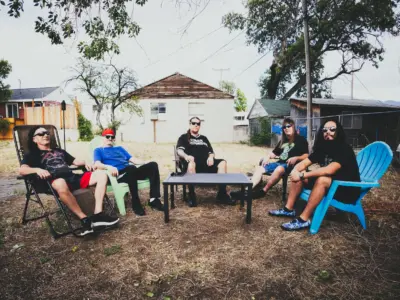 Five men sit in various types of lawn chairs on dead grass with a small foldable table in front of them, looking towards the camera.