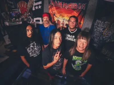 In a slight fisheye lens, five men stare up at the camera in a room covered in posters and tarps.