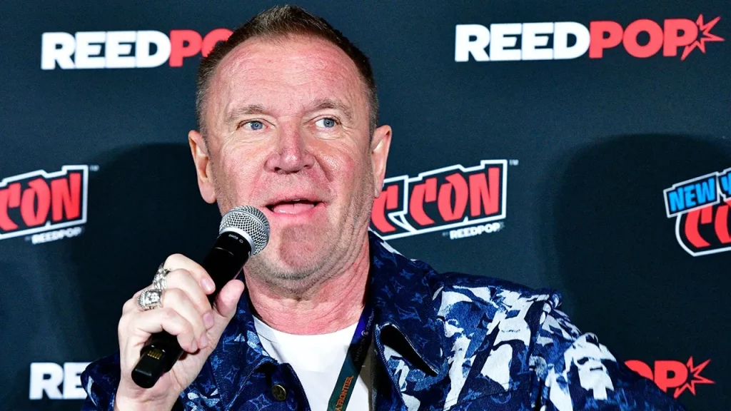 Renny Harlin stands in front of a convention step and repeat holds a microphone.