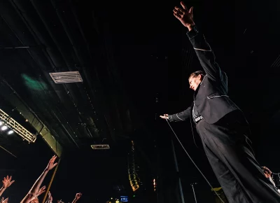 Pelle Almqvist of The Hives, stands near the edge of the stage and raises his hands as audience members hands raise to meet his.