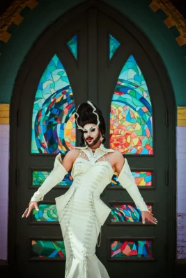 Salt Lake City based drag queen M'Lady Wood poses in a white dress infront of a church's stained glass windows.