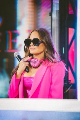 Meredith Marks holds a microphone. She wears a hot pink blazer and dress, bedazzled headphones and sunglasses.
