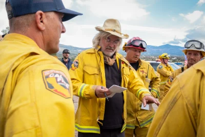 Paul Greengrass wearing a fireman's jacket, standing around other firemen.
