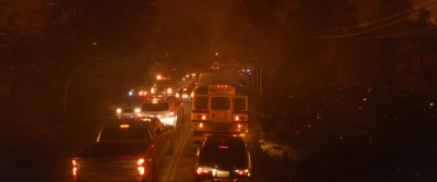 A herd of cars, including a school bus, drive down a smoky street.