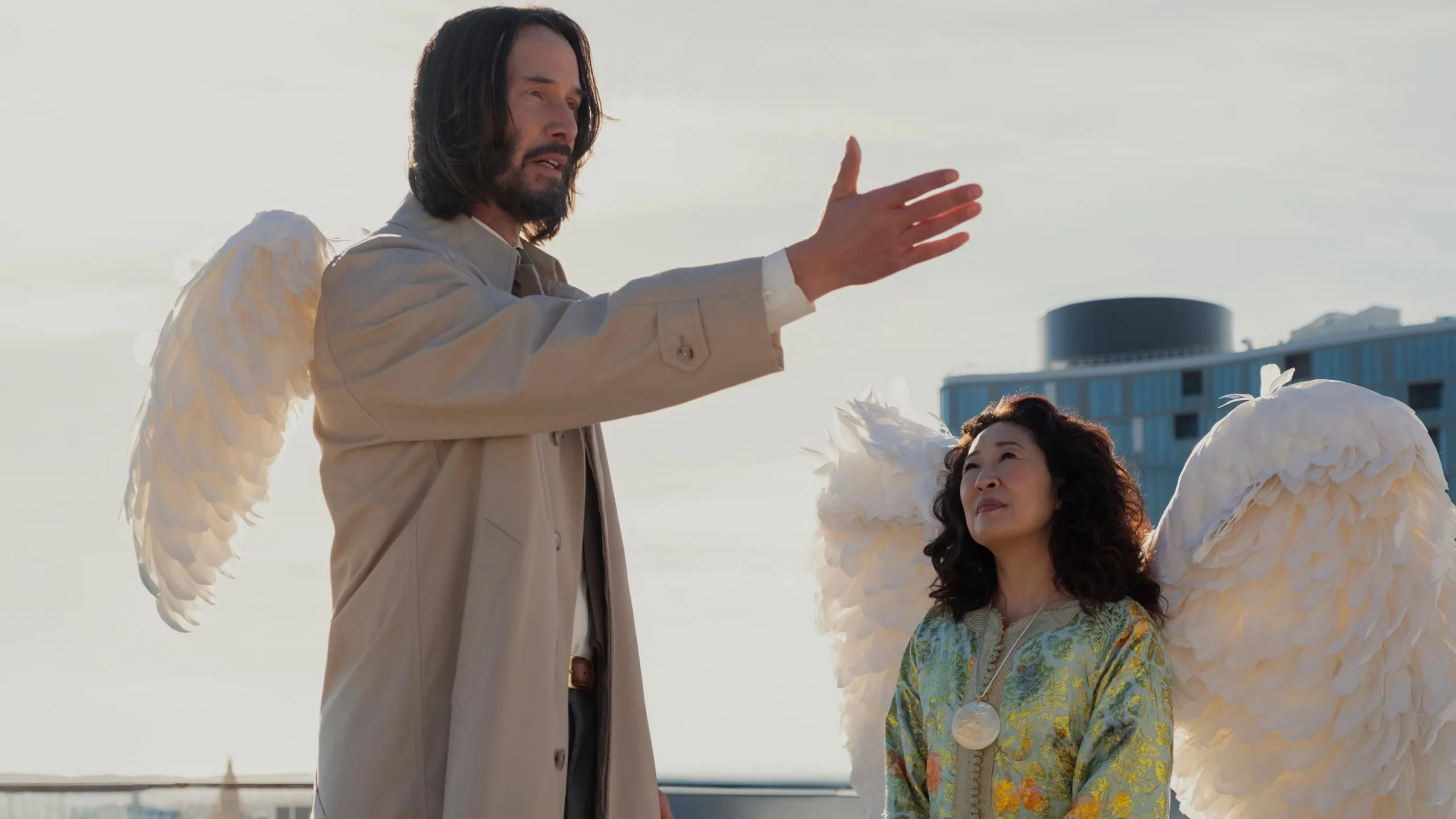 Keanu Reeves sticks his hand out wearing angel wings, as Sandra Oh also wears angel wings and looks up at him