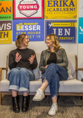 Finlayson and Morgan from Elevate sit talking on a couch. The wall behind them is covered in local election campaign signs from various candidates.