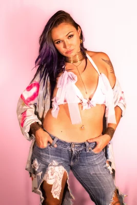 Katsi wearing a bra with laced wrapped around it and jeans, standing in front of a pink background
