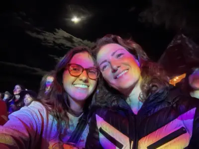 Two women cuddle together smiling under a full moon in rainbow light.